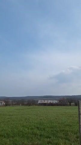 The video presents a static, wide-angle shot of a rural landscape on a partly cloudy day. In the foreground is a lush green field, likely grassland or pasture. Scattered along the field's edge are several concrete fence posts. The background features a row of leafless trees with a few buildings and a corrugated metal roof visible behind them. A low range of hills defines the horizon. The sky is blue with scattered white clouds, and a slight glare from the sun is visible in the upper right.