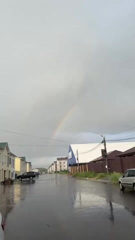 A rainbow arcs across a cloudy sky over a wet street. The street is lined with houses and a building with blue and white siding that has writing on it. There are cars parked along the street. The ground is wet and reflects the sky and buildings.