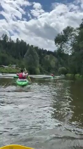 Here is a detailed description of the video:

The video shows people kayaking on a river. There are several kayaks visible, each with one or more occupants. The people in the kayaks are wearing life vests. They are paddling their kayaks along the river, moving in the same direction. The camera seems to be positioned from within one of the kayaks, capturing the scene from the paddler's perspective. The paddle of the kayak in which the cameraman is riding is visible in the foreground.

The river is surrounded by lush greenery, with trees and vegetation lining the banks. The sky is partly cloudy. The scene appears to be taken on a sunny day.
