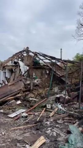 Okay, here's a forensic description of the video:

**Scene:** The video depicts a scene of devastation and destruction. A building is largely collapsed and reduced to a pile of charred and broken debris.

**Environment:** The environment appears to be an outdoor area, possibly a residential or industrial zone. The sky is overcast, suggesting a cloudy day. There are trees in the background, but they appear bare, indicating it might be late fall or early spring.

**Objects:** The scene is dominated by the wreckage of the building. This includes:

*   A collapsed roof structure, with exposed beams.
*   Scattered pieces of wood, metal, and other building materials.
*   Burned and charred debris, indicating a fire may have been involved.
*   Parts of a vehicle, including tires and a car hood, are visible amidst the wreckage.

**Overall Impression:** The video conveys a sense of destruction and loss. The extensive damage suggests a major incident, such as a fire, explosion, or natural disaster.