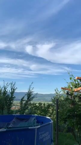 The video shows a landscape with a blue round swimming pool in the foreground, a lush green valley with a village visible in the middle distance, and mountains on the horizon under a bright blue sky with streaky white clouds. The swimming pool is partially covered. Trees and vegetation surround the pool area, with some of the branches moving gently in what seems to be a light breeze. The overall impression is that of a sunny, pleasant summer day.