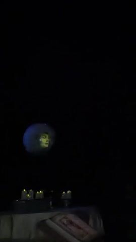 A video captured in a dimly lit room shows a disembodied face projected onto a spherical object that floats in the center of the frame. The face is that of a woman with yellow-toned skin, dark hair, and green eyes. She is surrounded by a blue, hazy aura. Below the projected face, a table is visible, draped with a white cloth. On the table, several unlit candles in holders are arranged. To the right of the floating face, another projection appears, this one appearing as a blue, ethereal figure or object. The room is predominantly dark, with the projected images and the faint glow of the candles being the only sources of light. The video appears to be filmed from a low angle, looking upwards towards the floating projection.
