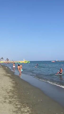 A wide shot captures a sandy beach with a clear blue sky overhead. Gentle waves lap at the shore, where several people are enjoying the water and sand. In the foreground, two women in bikinis stand near the water's edge, one appearing to be playing with a ball. To the right, a man is visible in the water, holding a paddleboard. Further out in the ocean, other beachgoers are swimming and engaging in various water activities. In the distance, a large, inflatable water park can be seen, adding to the lively atmosphere of the beach. The sun is shining brightly, indicating a warm and pleasant day.