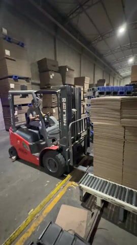 In a warehouse, a man operates a red and black forklift to move a large stack of cardboard sheets. The forklift is positioned next to a conveyor belt with rollers. The man, wearing a light-colored shirt, sits in the driver's seat, focused on maneuvering the forklift. The warehouse is filled with stacks of cardboard and blue plastic crates, and is well-lit by overhead lights. Yellow safety lines are painted on the floor to guide movement. The forklift lifts the stack of cardboard, preparing to transport it. The camera captures the scene from a stationary position.