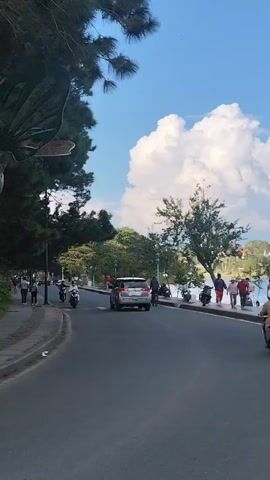A paved road curves to the left, bordered on the right by a body of water. Several people walk along the water's edge, and a few motorcycles are parked and in motion on the road and sidewalk. A white SUV is driving down the road. Trees line the sidewalk and overhang the road from the left side of the frame. A large, white cloud dominates the bright blue sky.