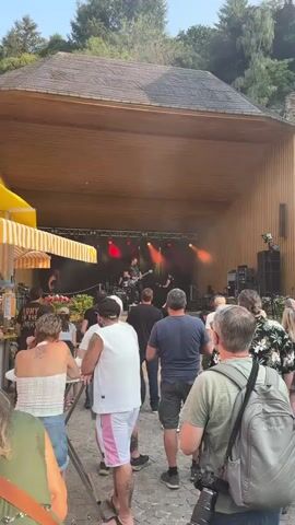A live band performs on an outdoor stage with a wooden roof. The band consists of a male guitarist in the foreground, a male drummer behind him, and another male guitarist on the right. A female figure with long hair is partially visible behind the drummer. The stage is lit with red and yellow spotlights. A crowd of people watches the performance from the area in front of the stage, with some standing and others potentially sitting at tables obscured from view. The overall environment appears to be a festival or outdoor concert setting.