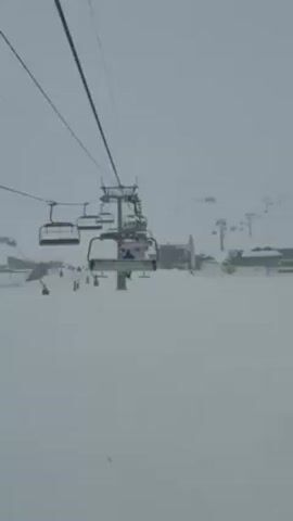 The video shows a ski lift moving over a snowy landscape. There are other skiers and snowboarders visible on the slope below, as well as buildings and construction equipment covered in snow. The weather appears overcast and snowy.