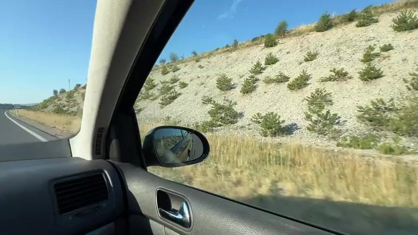 The video shows the interior of a car driving on a highway on a clear, sunny day. The perspective is from the passenger seat looking out the side window and through the windshield. The right side of the road features a steep, rocky embankment sparsely populated with small evergreen trees. The highway has multiple lanes and is bordered by a guardrail on the left. In the distance, a signpost is visible with directional arrows and text. The dashboard of the car is visible in the foreground, showing various buttons and air vents. A side mirror reflects the road behind the car. The sky is a bright, clear blue.