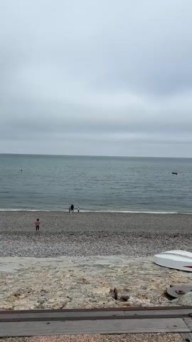 A slightly overcast day at a pebble beach. The sea is calm with small waves. In the foreground, a small white boat is overturned on the shore. Further back, on the pebble beach, are several people. Two adults and two children are near the water's edge, appearing to watch something in the sea. One child is running towards the water. Another child, wearing a pink top, is sitting on the beach to the left of the frame. A red buoy is visible in the sea in the distance. The overall scene is one of casual outdoor activity.