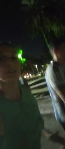Here's a detailed description of the video:

The video is taken at night, seemingly in a park or public outdoor space, based on the presence of trees and ground. The camera movement is shaky, suggesting it is handheld and the person is moving. The light source is primarily artificial lighting, such as street lamps, causing glare on the lens at times.

The primary subject is a woman wearing a green tank top. Her hair is pulled back. The video focuses on her as she moves and talks, although the audio isn't clear. 

Initially, another person, possibly male, is briefly visible in the background wearing a white shirt with light blue stripes. However, the focus quickly shifts to the woman in the foreground. The ground appears to be a light-colored surface, possibly concrete or packed dirt.