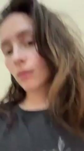 A woman with long, brown, somewhat messy hair is centered in the frame against a light-colored wall. She is wearing a gray sleeveless shirt. She looks down briefly, then straight ahead, then up towards the left. She appears to be listening intently to something. There is music playing in the background.