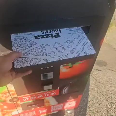 A hand is shown holding a pizza box with a pattern of pizza slices and utensils on it. The box is being lowered into a dark opening of a black machine, possibly a vending machine or delivery locker. The machine has text visible that says "MINUTES". Below the opening, there is a glimpse of a red object, and further down, a label with a phone number "07 85 58 16 40" is visible on another red object. The pizza inside the box appears to be topped with meat and a drizzle of sauce. The scene is outdoors, with a paved surface and greenery visible in the background, suggesting a sunny day.