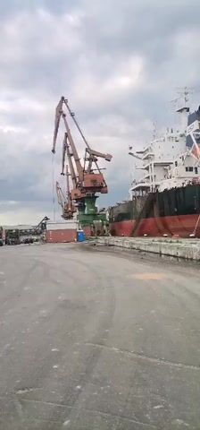 The video shows a large cargo ship named “CARAVOS HARMONY” docked at a port. The ship is black and red with a white upper deck and a blue and white navigation tower. It’s secured to the concrete pier with ropes. Several large, rusty cranes are positioned next to the ship, presumably for loading and unloading cargo. A few shipping containers are also visible. The sky is overcast with grey clouds, and the pavement is damp, suggesting recent rain.  A seagull flies in the background. The scene appears to be taken at eye-level from a slightly moving position on the pier.
