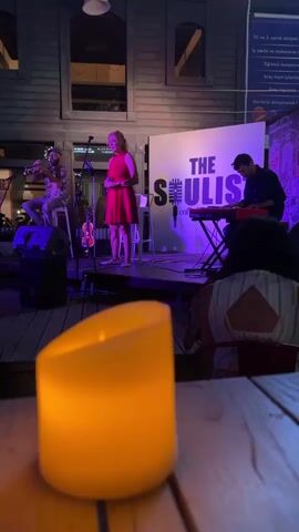 A live music performance is taking place indoors at night, with stage lighting creating a purple and red ambiance. In the foreground, a lit yellow candle is on a wooden table, out of focus. Behind the candle, a band performs on a raised stage. A woman in a red dress stands center stage, holding a microphone and singing. To her left, a man plays the trumpet while seated. To her right, a man plays a keyboard. A banner behind the performers reads "THE SOULIST." In the audience, a person with dark curly hair is visible, facing away from the camera. The background appears to be a wooden building with windows. A sign with text in Turkish is visible on the far right.