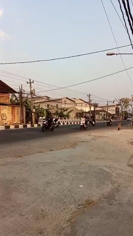 A clear, bright sky is visible above a street scene. The street is paved with asphalt and has a concrete shoulder. Several motorcycles with riders are moving along the street. A silver car is also present. Buildings line one side of the street, and there are utility poles and overhead wires. The lighting suggests it is either early morning or late afternoon.