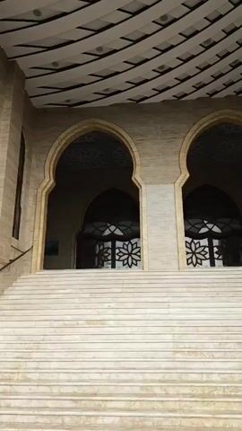 The video shows the exterior of a modern mosque. The camera pans up the stairs leading to the entrance. The mosque has a light-colored stone facade and arched doorways. The ceiling above the entrance is decorated with a geometric pattern. To the right, a tall minaret with multiple levels and windows rises into a blue sky with white clouds. The minaret also appears to be constructed of light-colored stone. Trees and utility poles are visible in the background. There are no people visible in the video.