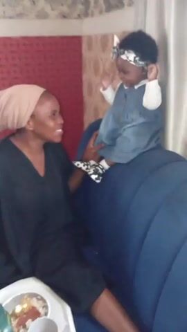 A woman wearing a black dress and a light brown headscarf sits on a blue sofa. In front of her is a baby standing on the back of the sofa, wearing a denim dress, a white long-sleeved shirt, polka dot shoes, and a bow headband. The woman looks up and smiles at the baby. There's a white tray with food on it in front of the woman. The background includes a patterned wall and white curtains.