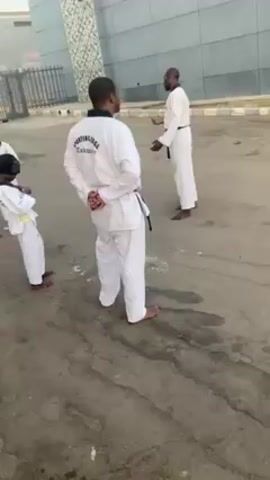 The video shows a Taekwondo instructor in a white uniform and black belt teaching students outdoors. The instructor demonstrates a kick while the students, also in white uniforms and various belt colors, observe. The scene is set in a parking lot or similar paved area next to a modern building. The students are barefoot.