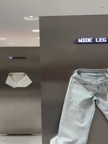 The media file displays a retail environment, likely a clothing store, with a focus on denim. Two items of clothing are prominently featured: a pair of white denim mini shorts hanging on a wall to the left, and a pair of light blue wide-leg jeans draped over a counter or display to the right.

Above the mini shorts, a digital sign displays the text "MINI SHORTS". To the right of this, another digital sign cycles through different text. Initially, it shows "DE LEG", then transitions to "MINI", and finally to "WIDE LEG".

The setting appears to be indoors, with bright, diffused lighting from ceiling fixtures. The walls are a sleek, metallic gray, and the floor visible at the bottom is a neutral tiled surface. There are no people visible in the frame, and no indication of the time of day or weather conditions outside. The scene is clean and minimalist, characteristic of a modern retail display. The focus is solely on the merchandise and the informative digital signage. The location context is Aldaia, Spain, suggesting this is a store within that region.