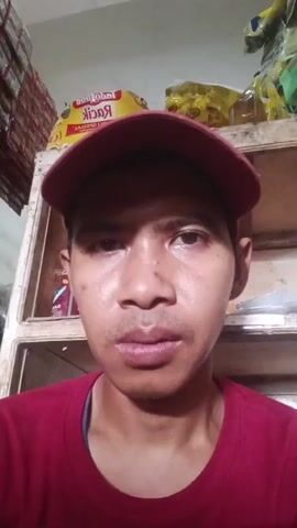 A person wearing a red t-shirt and a red baseball cap looks into the camera. They appear to be in a store or warehouse, with shelves filled with various products behind them. The shelves are wooden and stacked with bags and boxes of goods, including a yellow bag with red lettering that says "IndoFood." The lighting is dim, suggesting indoor lighting. The person's face is close to the camera, and their expression is neutral. The video appears to be a selfie-style recording.
