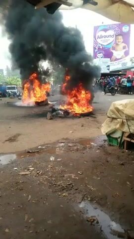 The video depicts an outdoor scene in Ndenderu ward, Kenya, during daylight hours, characterized by an ongoing protest. Multiple tires are burning on a dirt road, producing large, intense orange flames and thick, dark black smoke that billows upwards.

A crowd of people, primarily men, are gathered in the background, observing the burning tires. Some individuals can be seen standing near the fires, and at one point, a man in a brown jacket and a red cap walks past the burning tires.

The ground around the fires is unpaved, covered in dirt and scattered with rocks and debris. In the background, there are commercial structures, including market stalls covered with beige tarps. Several vehicles are visible further down the road.

Two large billboards stand out in the background: one advertising "ALLROUND REDEFINES PANTS" with the text "FREE TO STRETCH OT FALL OFF" and an image of a baby, and another promoting "MASS VOTER REGISTRATION" featuring "HON. ALICE NG'ANG'A #MamaSimba" with the slogan "Kura Yako, Future Yako!". The sky appears overcast, partly obscured by the heavy smoke from the fires.