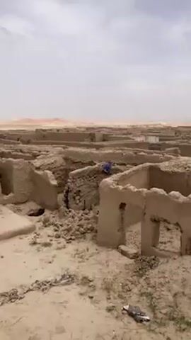 The video presents an extensive panorama of an abandoned settlement, identified as Saranan, Pakistan. The scene is dominated by numerous ruined mud-brick structures, appearing as low walls and foundations of what were once buildings, stretching far into the arid landscape. The ground is dry, sandy, and strewn with debris, including a white plastic bag and some very sparse green vegetation. One structure features a section of corrugated metal sheeting, painted blue along its lower edge. The sky is overcast with light clouds, and distant hills or mountains are visible on the horizon. No people or specific activities are visible; the video simply pans across the desolate ruins under daylight conditions.