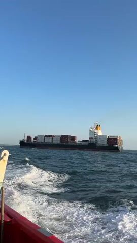 A container ship travels across a wide expanse of choppy, blue ocean under a clear, bright blue sky. The ship is viewed from the stern of a smaller vessel, the red side of which is visible in the foreground. The container ship is laden with stacked shipping containers, most of which are red. A plume of dark smoke rises from the ship's stack. The smaller vessel is creating a white wake as it moves through the water.