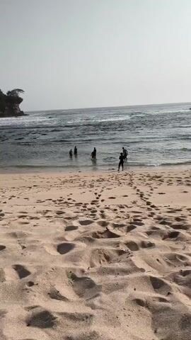 The video shows a sandy beach with footprints in the foreground. In the mid-ground, several people are standing in the shallow water of the ocean. One person is visible on the beach in the mid-ground, facing away from the camera. In the background, there are waves and a rock formation with vegetation on the left. The sky is overcast.