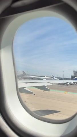 From the interior of an airplane, the view through the window reveals the wing of the plane, the airport tarmac, and another plane, an Etihad Airways plane. The sky is blue with thin, streaky clouds. A small patch of weeds is visible on the tarmac. The window frame is white.  Some voices are heard.