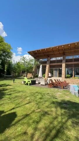 This video displays a luxurious outdoor scene with a modern house and a swimming pool. The house features a distinctive architectural design with a sloping wooden roof supported by stone pillars. Large glass windows offer views of the interior and the surrounding landscape. In the foreground, a lush green lawn is visible, with shadows cast from nearby trees. The lawn is dotted with outdoor furniture, including lounge chairs and a table, suggesting a recreational area. To the right of the house, a bright blue swimming pool reflects the clear blue sky. Surrounding the pool is a neatly manicured hedge, adding to the well-maintained aesthetic of the property. The overall impression is one of a serene and upscale residential setting on a sunny day.