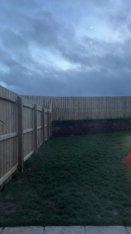 The video shows a backyard with a lawn and a wooden fence on two sides. The fence is made of vertical planks and appears to be relatively new. Along the back fence, there's a low brick retaining wall. A green garden hose is attached to the left fence. The grass is neatly cut. The sky is cloudy and overcast, suggesting it is either early morning or late evening. The lighting is dim due to the cloud cover. There are no people visible in the video.