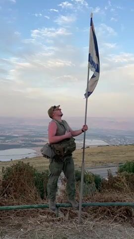 Here is a description of the video. 

The video shows a man standing on top of a hill holding an Israeli flag. The man is wearing a green vest and pants and has a backpack. He is standing on a metal railing overlooking a valley with a lake in the distance. The sky is blue with some clouds. The man is smiling and looking at the camera.