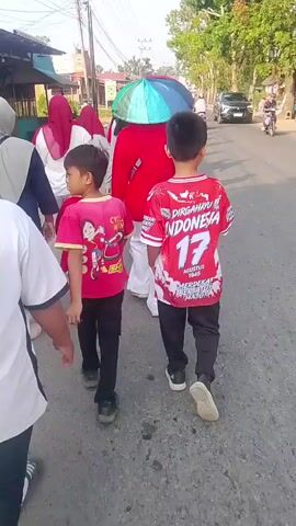 The video shows a group of people walking down a paved road, possibly in a village or suburban area. The time appears to be daytime, with bright sunlight. The group consists of adults and children, some of whom are wearing red and white clothing, suggesting a celebration or national event. One person in the group is holding a colorful umbrella. There are buildings and trees visible along the sides of the road, and a few motorcycles and cars are present, indicating traffic. The camera is positioned behind the group, capturing their movement from a rear perspective. The overall atmosphere seems to be one of a casual outing or procession.