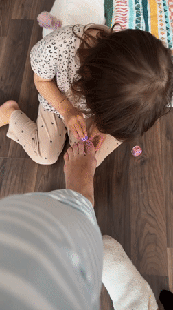 Child gives adult pink pedicure on wooden floor