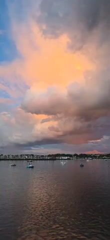 The camera captures a scenic view of a tranquil body of water, dotted with several boats bobbing gently on the surface. The opposite shore is lined with a cityscape, possibly a town or a collection of buildings, extending along the horizon. The most striking feature of the scene is the sky, filled with dramatic, voluminous clouds. The clouds are illuminated with hues of orange, pink, and gray, suggesting a vibrant sunrise or sunset. The color palette of the sky is mirrored in the water's reflection, adding to the overall picturesque quality of the scene. The video appears to be still or very slowly panning.
