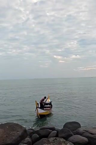 The scene captures a person in a small yellow and white boat on the sea. The person, wearing a dark shirt, is crouched down inside the boat, seemingly working or adjusting something. The boat is positioned near a rocky shoreline, with several large, dark rocks visible in the foreground. The sea stretches out to the horizon, and the sky is overcast with a layer of mottled clouds. The overall lighting suggests it's either early morning or late afternoon.