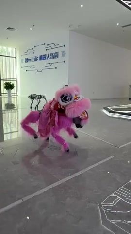 The video shows a child operating a small, robotic, pink lion dance puppet inside a modern, brightly lit building. The puppet, adorned with a traditional lion dance head and pink fur, moves under the child's control via a handheld remote. The child, wearing a white t-shirt and blue jeans, stands facing the puppet, his attention focused on the remote and its actions. There are other people in the background, partially visible, watching the demonstration. The floor is made of polished tile and has visible line designs, and the walls are white. In the background, there is a sign written in Chinese. In addition, there are informational kiosks showing the words “MagicDog-W” and “Unitree Go2”.