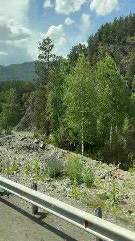The video shows a view from a vehicle moving along a road next to a river. The river is wide and has a turquoise color. It flows through a mountainous, forested area. The sky is blue with scattered white clouds. The foreground includes a guardrail and rocky terrain with some vegetation. Trees, including pines and birches, are visible along the riverbank and on the hillsides. The camera perspective suggests the video was recorded from a moving car or bus. There are no people visible in the video. The environment appears natural and somewhat wild.