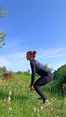 A woman with red hair pulled up in a bun is performing jump squats outdoors in a grassy field under a clear blue sky. She is wearing black leggings and a black long-sleeved shirt with a geometric pattern on the shoulders and sleeves. She is captured mid-jump and then in a squatting position. The background consists of green grass, some wildflowers, and trees.