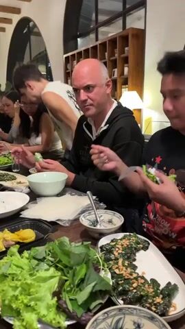 The video shows a group of people sitting around a table, preparing and eating a meal. The table is laden with various dishes, including fresh greens, wrapped rolls, and a plate of cooked vegetables topped with nuts. The people are engaged in the act of preparing their food, with some wrapping ingredients in rice paper and others holding their prepared rolls. The environment appears to be a casual dining setting, possibly a restaurant, with warm lighting and wooden furniture. The people seem to be enjoying their meal and each other's company. The focus is on the food and the interactive process of preparing and consuming it.