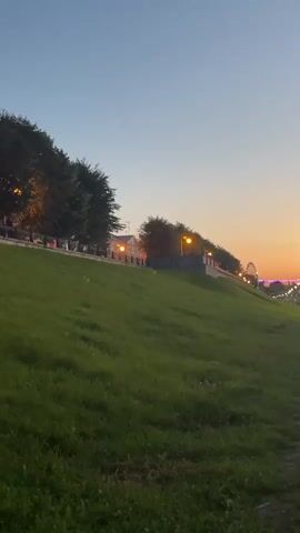 A short video shot at dusk from a low angle, looking up a grassy hill towards a row of trees and streetlights. The sky is a gradient of blue and orange. Beyond the trees and lights, a Ferris wheel and other city lights are visible. A paved path runs alongside the hill on the right side of the frame. The lighting suggests it is twilight or early evening.