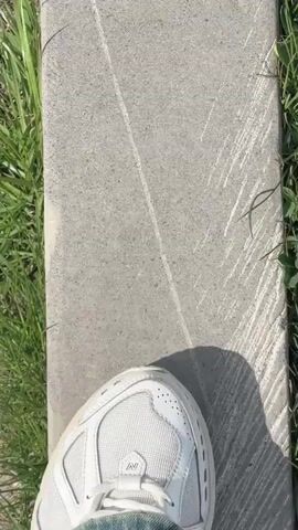 The video begins with a close-up of a concrete curb with white markings and some grass growing next to it. The view then pans to show the curb extending into the distance, bordered by grass and a dirt path. A cigarette butt is visible on the path. The scene transitions to a wider shot of a group of people walking along a paved path in a mountainous area. There are small buildings and parked cars visible in the background. The video then focuses on the majestic, snow-capped mountains under a cloudy sky. The last shot shows a close-up of the clouds and the peaks of the mountains.