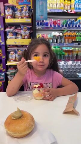 A young girl with brown hair in pigtails is sitting at a white table, eating fruit salad from a plastic cup with a wooden spoon. She is wearing a pink t-shirt. In front of her is a donut with green frosting. Behind her is a store aisle with shelves stocked with snacks and drinks. The girl is looking at the camera and chewing the fruit.