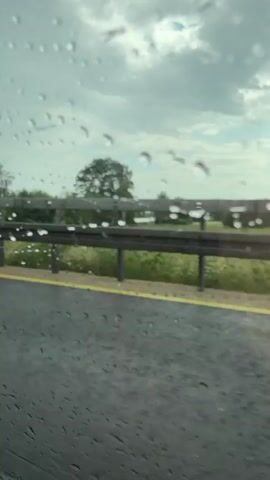 The video shows a view through a rain-streaked window, likely from inside a vehicle. The scene outside is a highway with a guardrail and trees lining the side. The road surface appears wet, indicating it is raining. A silver car passes by, with its license plate visible. The sky is overcast with clouds. The perspective is low, suggesting the camera is positioned near the ground or at the height of a car's window. There are no people visible in the frame. The overall impression is a drive on a wet, cloudy day.
