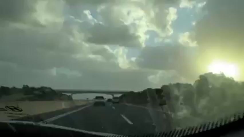 The view from inside a car driving on a highway captures a partly cloudy sky with the sun shining brightly. A bridge passes overhead, and other cars are visible on the road. The landscape includes trees and hills. The overall impression is of a highway drive on a day with mixed weather.