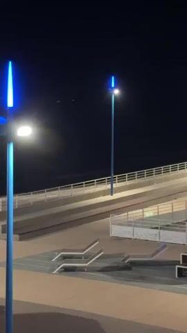 The video shows a nighttime scene with two blue lampposts. The lamppost on the left is closer to the camera and has a white light at the bottom and a blue light at the top. The lamppost on the right is further away and has a white light at the top, with a blue light above it. There is a ramp or elevated walkway in the background with railings. In the foreground, there are some modern-style benches or seating arrangements. The sky is dark and clear, suggesting it is nighttime. There is no visible movement or activity in the scene.