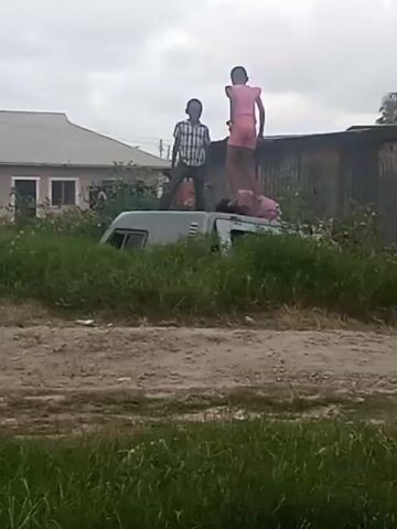 Two children are standing on top of a van in a grassy area. The van is gray and appears to be old. A third child is partially visible behind them on top of the van. The children are silhouetted against a cloudy sky. In the background, there is a house and a corrugated metal structure. The ground is dirt.