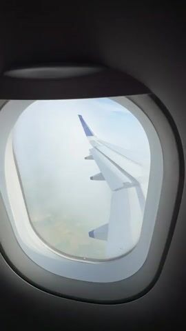 This is a first-person point-of-view video filmed from inside an airplane, looking out of the window. The airplane's wing, a large white structure with blue wingtips, dominates the right side of the frame. The wing appears to be in flight, as the background shows a landscape of fields and some clouds below. The window frame, a dark grey oval, encircles the view of the wing and the landscape. The sky is a mix of blue and white, with some fluffy clouds visible. The footage is slightly shaky, indicating movement, likely from the airplane in motion. The overall scene depicts a typical view from an airplane during flight.