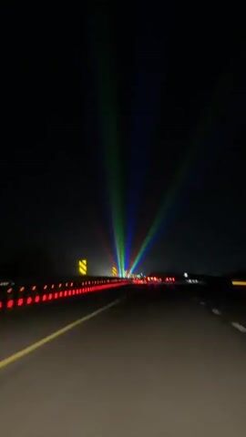 The video shows a view from the perspective of a person driving on a highway at night. The road ahead is illuminated by the headlights of other vehicles and by a set of colorful laser lights projected into the dark sky. The laser lights appear to be red, green, and blue, and they are fanned out, creating beams that reach high into the night. The highway is lined with red reflectors and traffic signs. The overall impression is one of a festive or celebratory light display happening along the roadside.