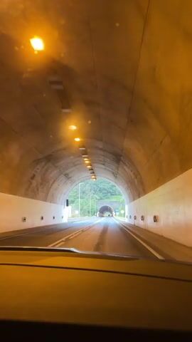 The video depicts a car driving through two tunnels.  The first shot is inside a brightly lit tunnel. The walls are grey concrete. The driver exits the tunnel and proceeds down a wet road toward a second, shorter tunnel surrounded by lush greenery and a cloudy sky. A sign to the right of the tunnel entrance indicates “Túnel da Madalena”.